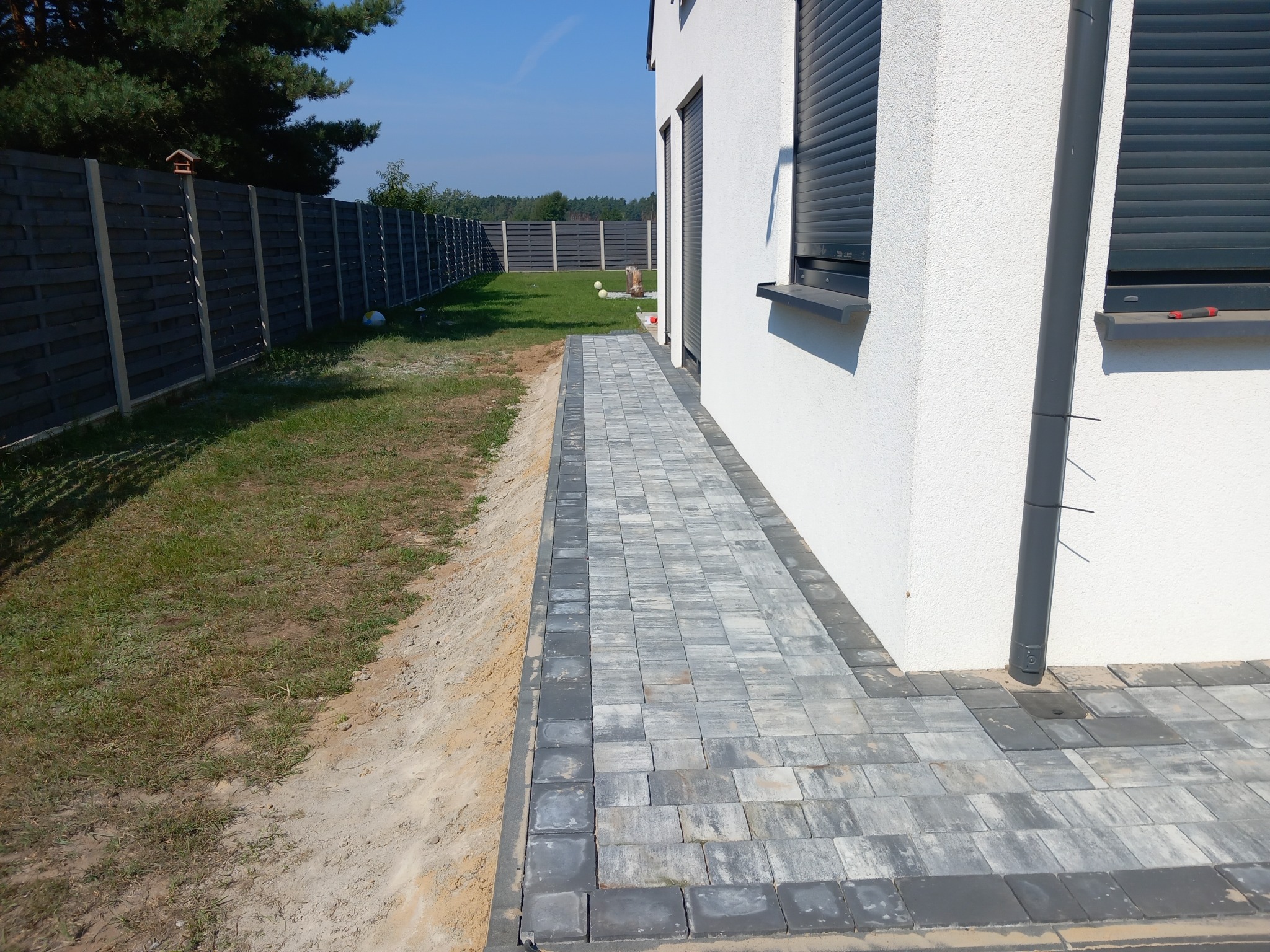 Ścieżka z szarej kostki brukowej prowadząca wzdłuż białego budynku z ciemnymi roletami, obok trawnik i ciemnoszare ogrodzenie panelowe.