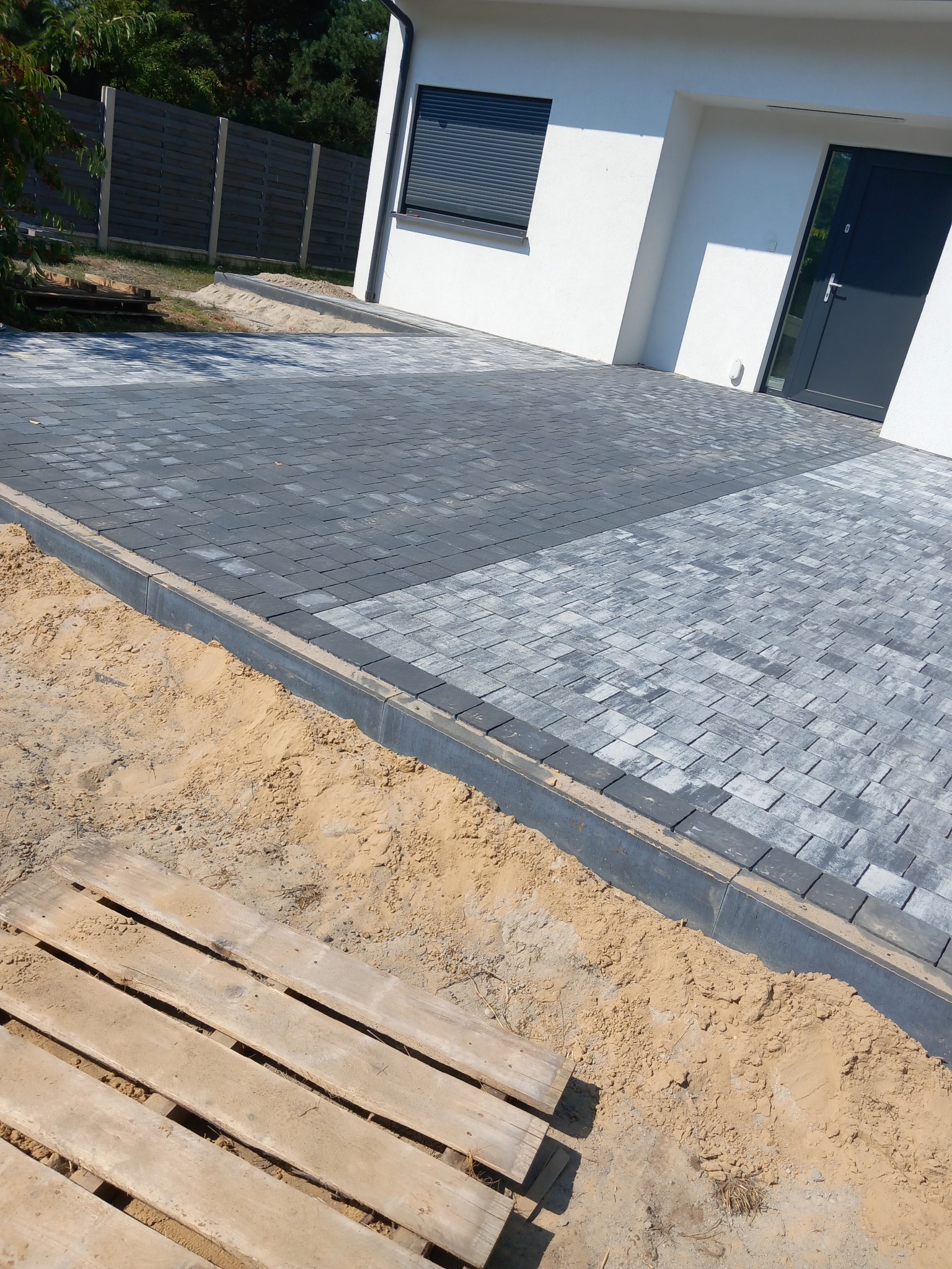 Ułożona dwukolorowa kostka brukowa przed nowoczesnym domem z szarą stolarką okienną i drzwiową, widoczna paleta i piasek na obrzeżach.