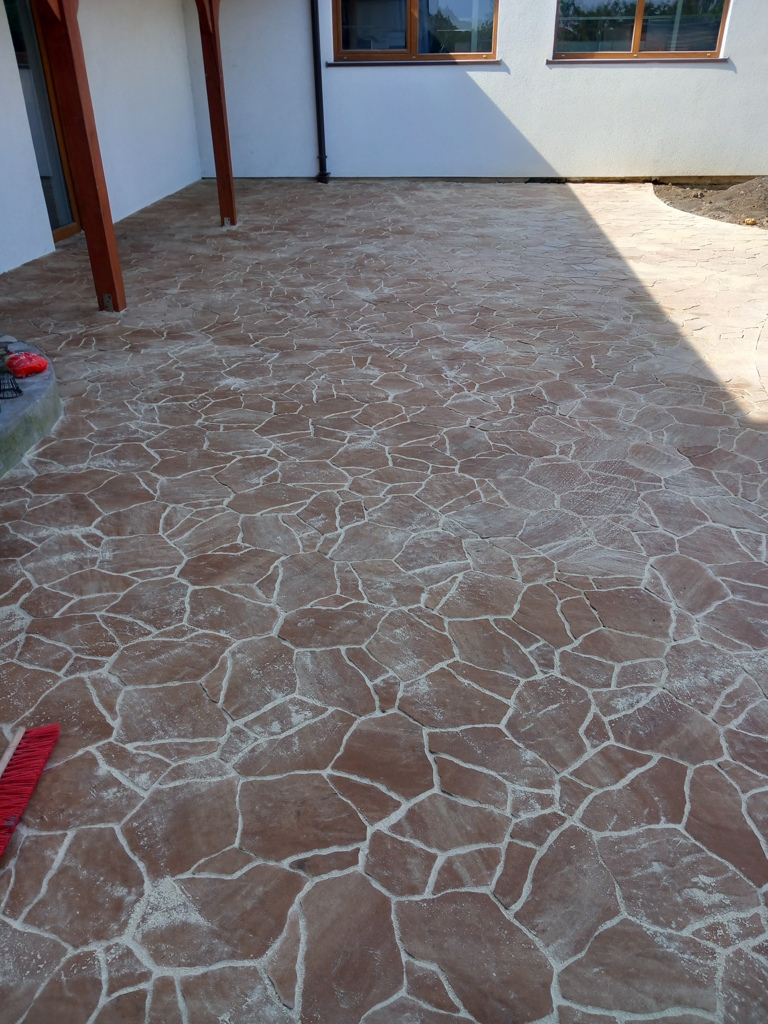 Nowa, brązowa nawierzchnia tarasu z nieregularnych płyt kamiennych z jasnymi fugami, częściowo zacieniona, widoczne fragmenty budynku i czerwona szczotka.