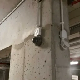 Kamera monitoringu kopułkowa zamontowana na surowej betonowej ścianie, obok czujnik ruchu i instalacja elektryczna w białych rurkach.