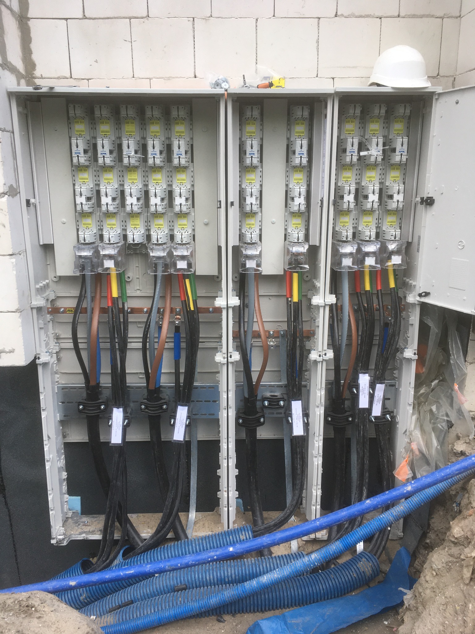 Otwarta szafa elektryczna z zainstalowanymi bezpiecznikami i podłączonymi kablami oznaczonymi kolorami, biały kask ochronny na górze, niebieskie rury karbowane na dole.