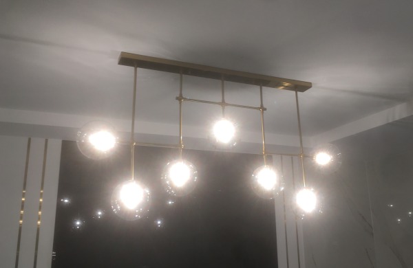 Nowoczesna lampa sufitowa z siedmioma szklanymi kloszami w kształcie kul, zawieszonymi na złotych, metalowych prętach, oświetlająca wnętrze z minimalistycznym wystrojem.