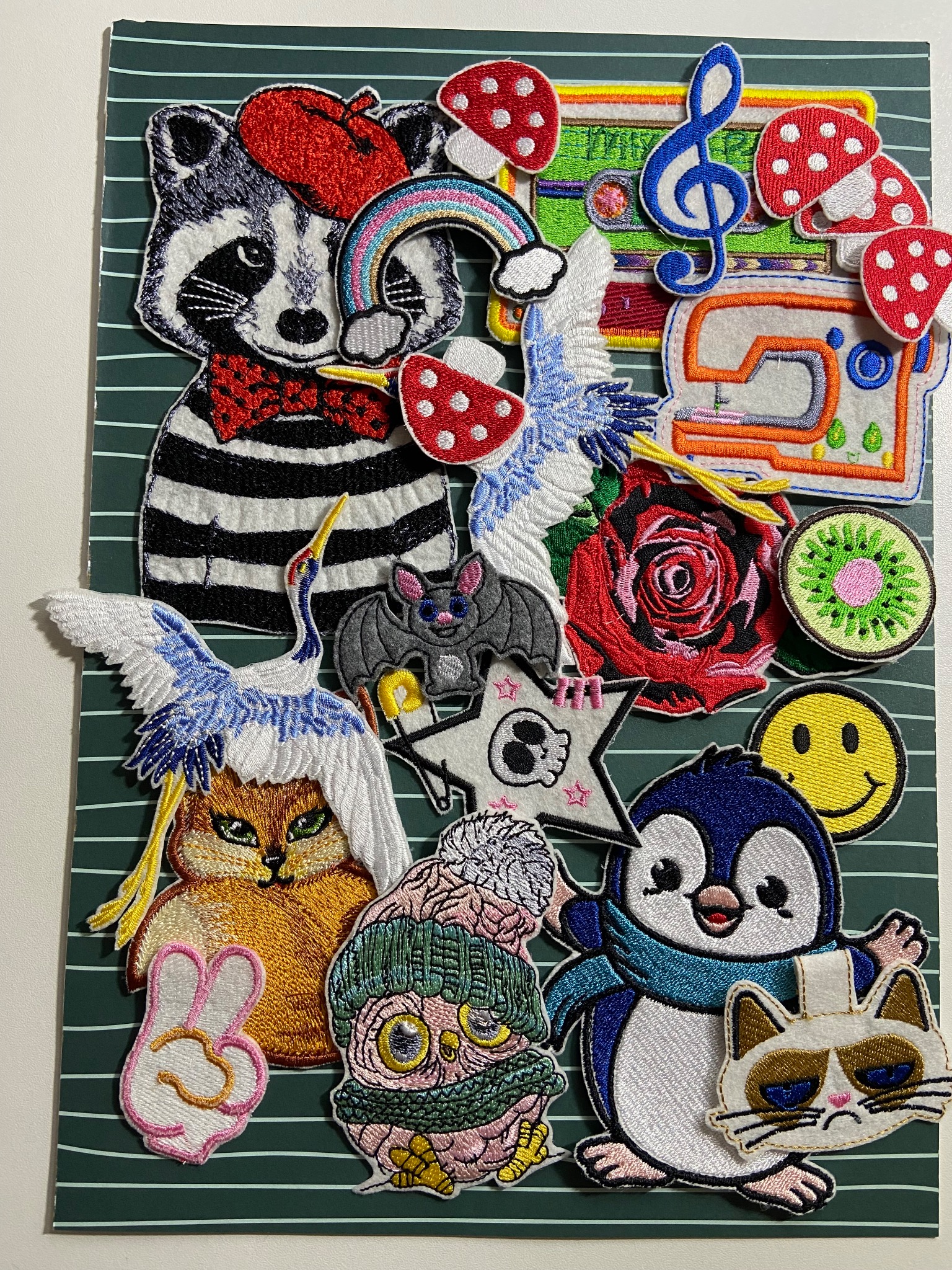 Kolekcja różnorodnych, kolorowych naszywek z motywami zwierzęcymi, roślinnymi i przedmiotami codziennego użytku, ułożonych na zielonym tle w paski. Widoczne m.in. szop w berecie, żuraw, nietoperz...