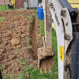 Mini koparka Bobcat w trakcie wykonywania wąskiego wykopu w ziemi, widok z kabiny operatora, w tle dom jednorodzinny i ogród.