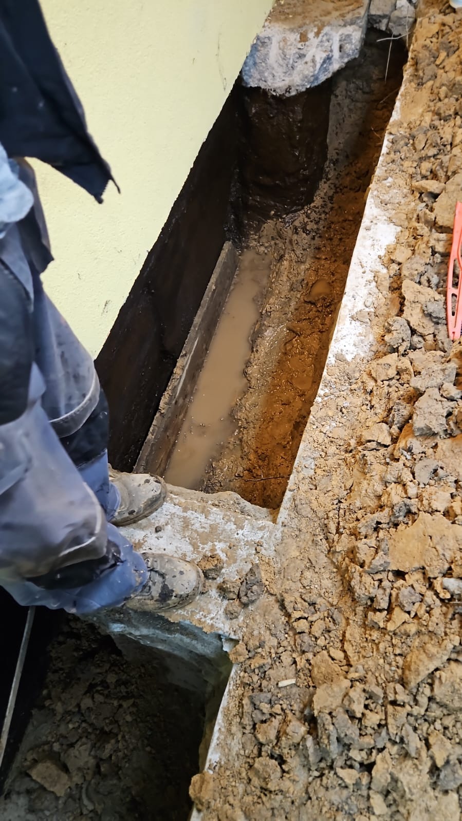 Wykop liniowy wypełniony wodą, widoczne fragmenty ściany budynku i buty robocze osoby stojącej obok.