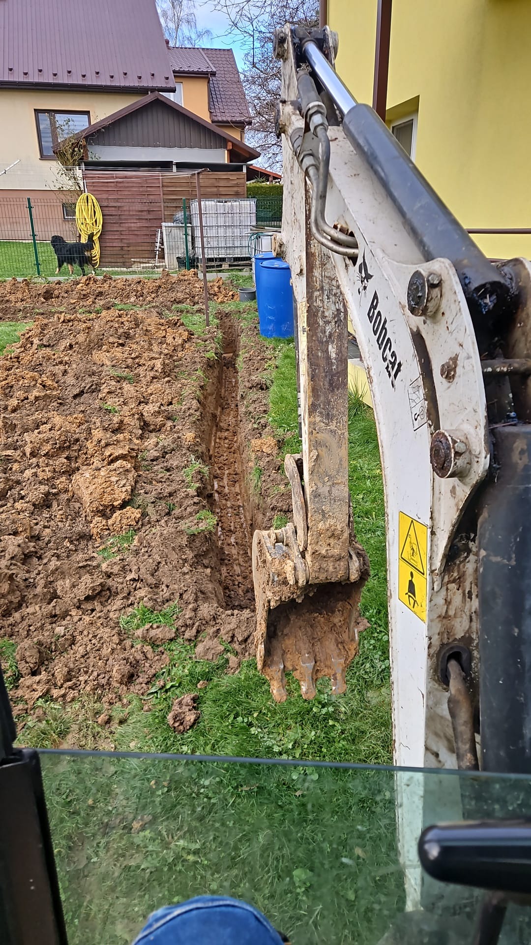 Mini koparka Bobcat w trakcie wykonywania wąskiego wykopu w ziemi, widok z kabiny operatora, w tle dom jednorodzinny i ogród.