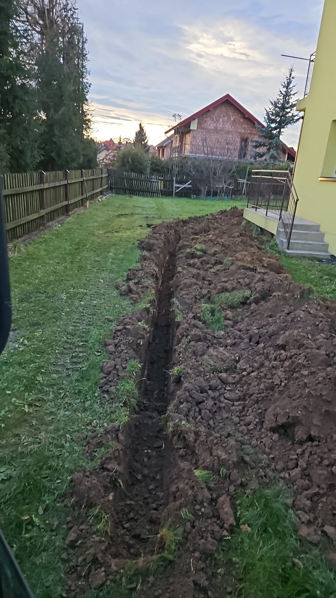Wykopany wąski rów w trawiastym ogrodzie, przygotowany pod instalacje, z widocznym budynkiem mieszkalnym w tle.