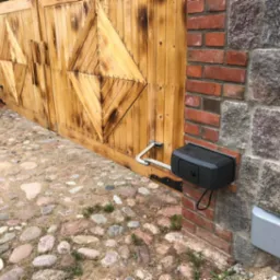 Drewniana brama wjazdowa z automatyką, widoczne siłowniki przytwierdzone do murowanego słupka z cegły i kamienia, oraz kamienny bruk podjazdu.