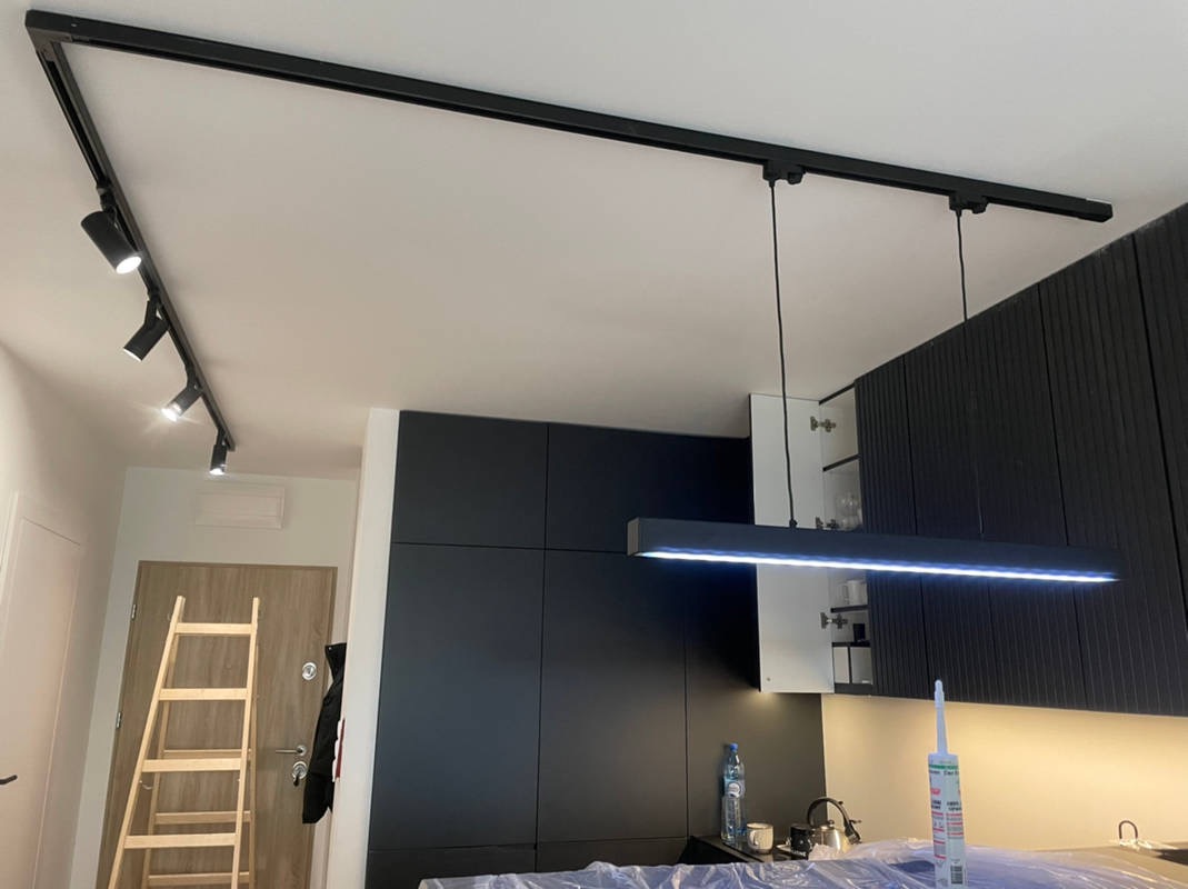 Czarna szyna oświetleniowa z reflektorami punktowymi i podłużna lampa LED wisząca na tle zabudowy kuchennej z czarnymi frontami. W tle drabina malarska.