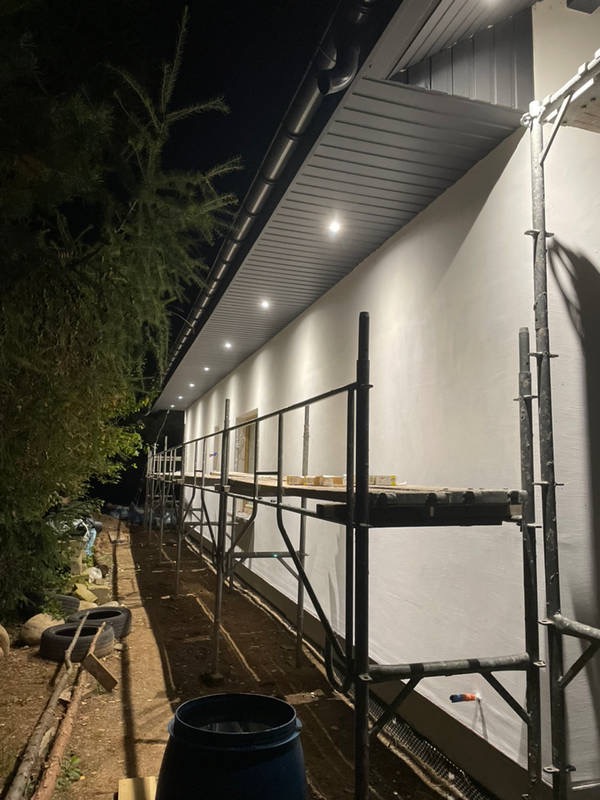 Elewacja budynku oświetlona szeregiem punktowych lamp podsufitowych, widoczne rusztowanie wzdłuż ściany w trakcie prac.