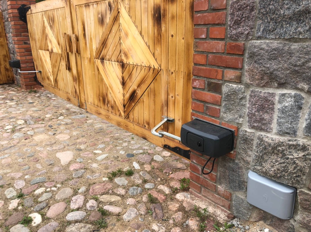 Drewniana brama wjazdowa z automatyką, widoczne siłowniki przytwierdzone do murowanego słupka z cegły i kamienia, oraz kamienny bruk podjazdu.