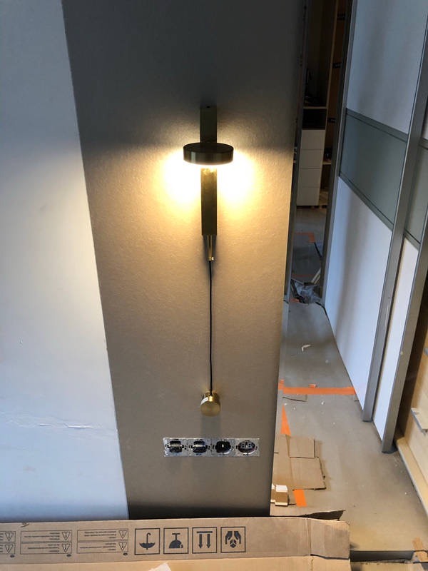 Zainstalowany, nowoczesny kinkiet w kolorze szczotkowanego złota, z widocznymi czterema gniazdkami elektrycznymi poniżej, zamontowany na ścianie w trakcie remontu.