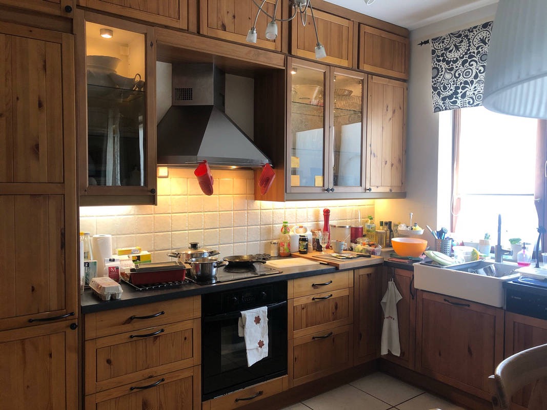 Kuchnia z drewnianymi szafkami, czarnym blatem i płytą grzewczą. Nad blatem zamontowany okap z czerwonymi rękawicami kuchennymi. Oświetlenie podszafkowe podkreśla fakturę płytek.