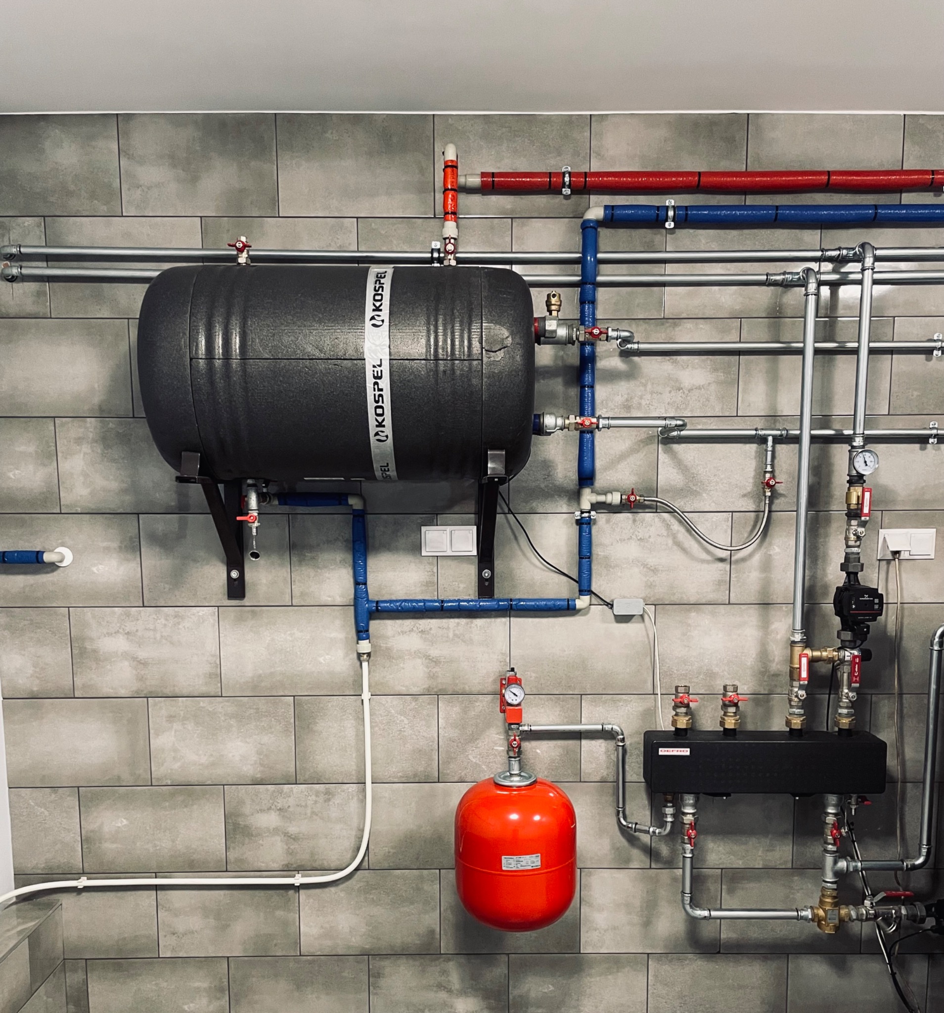 Szary akumulator ciepła Kospel zamontowany na ścianie z szarych płytek, z widocznymi rurami w kolorach niebieskim, czerwonym i srebrnym oraz elementami instalacji grzewczej, w tym czerwonym...
