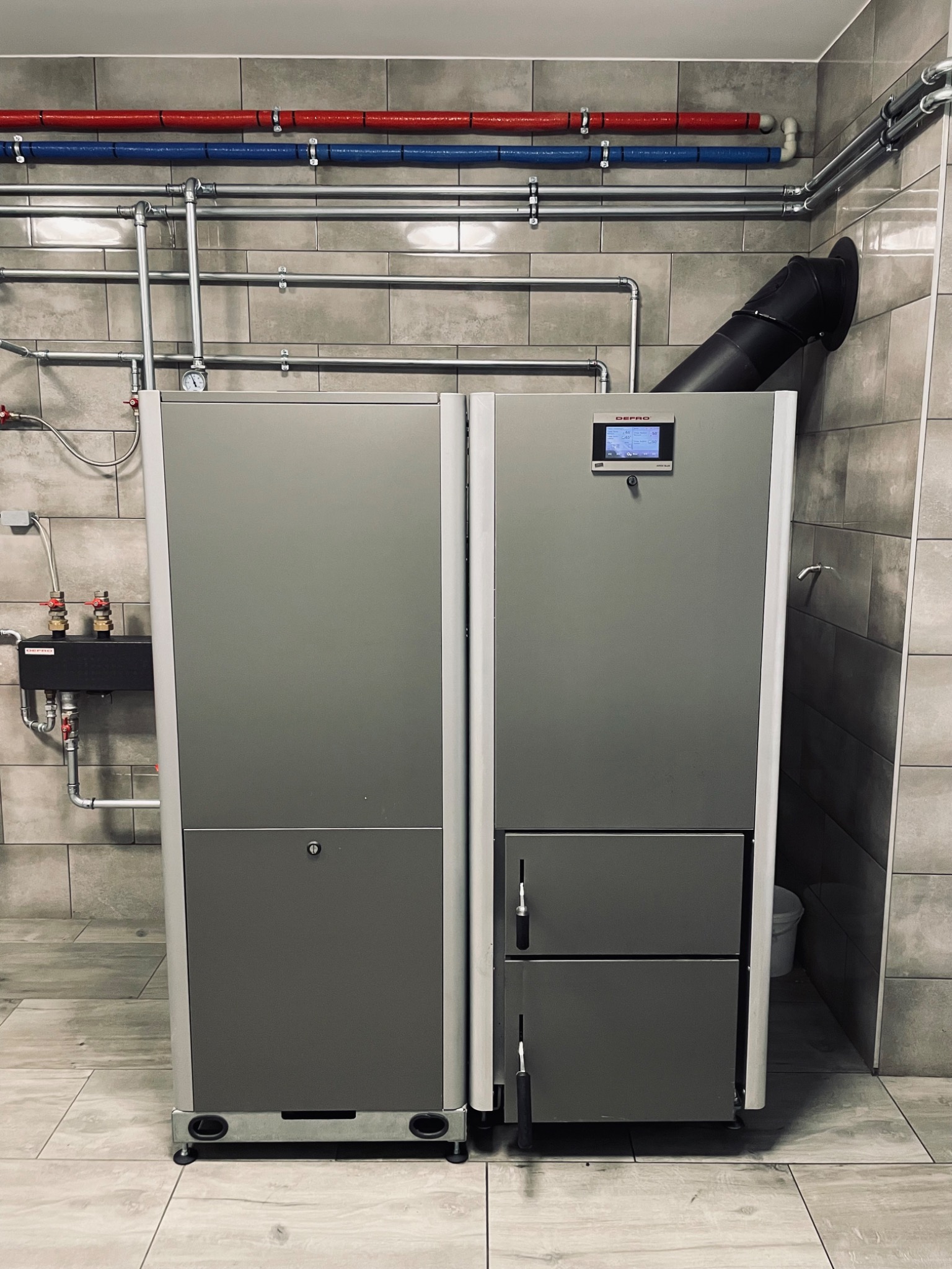 Szary piec z wyświetlaczem i kominem w pomieszczeniu z szarą, kafelkową ścianą i podłogą. Nad piecem widoczne rury w izolacji czerwonej i niebieskiej.