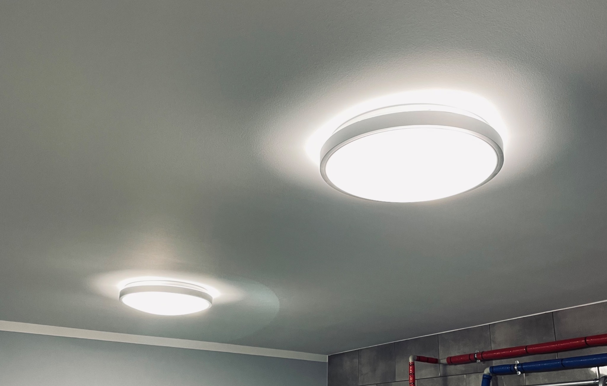 Dwa okrągłe, białe plafony LED zamontowane na szarym suficie w łazience z szarymi płytkami i widocznymi rurami instalacji wodnej.