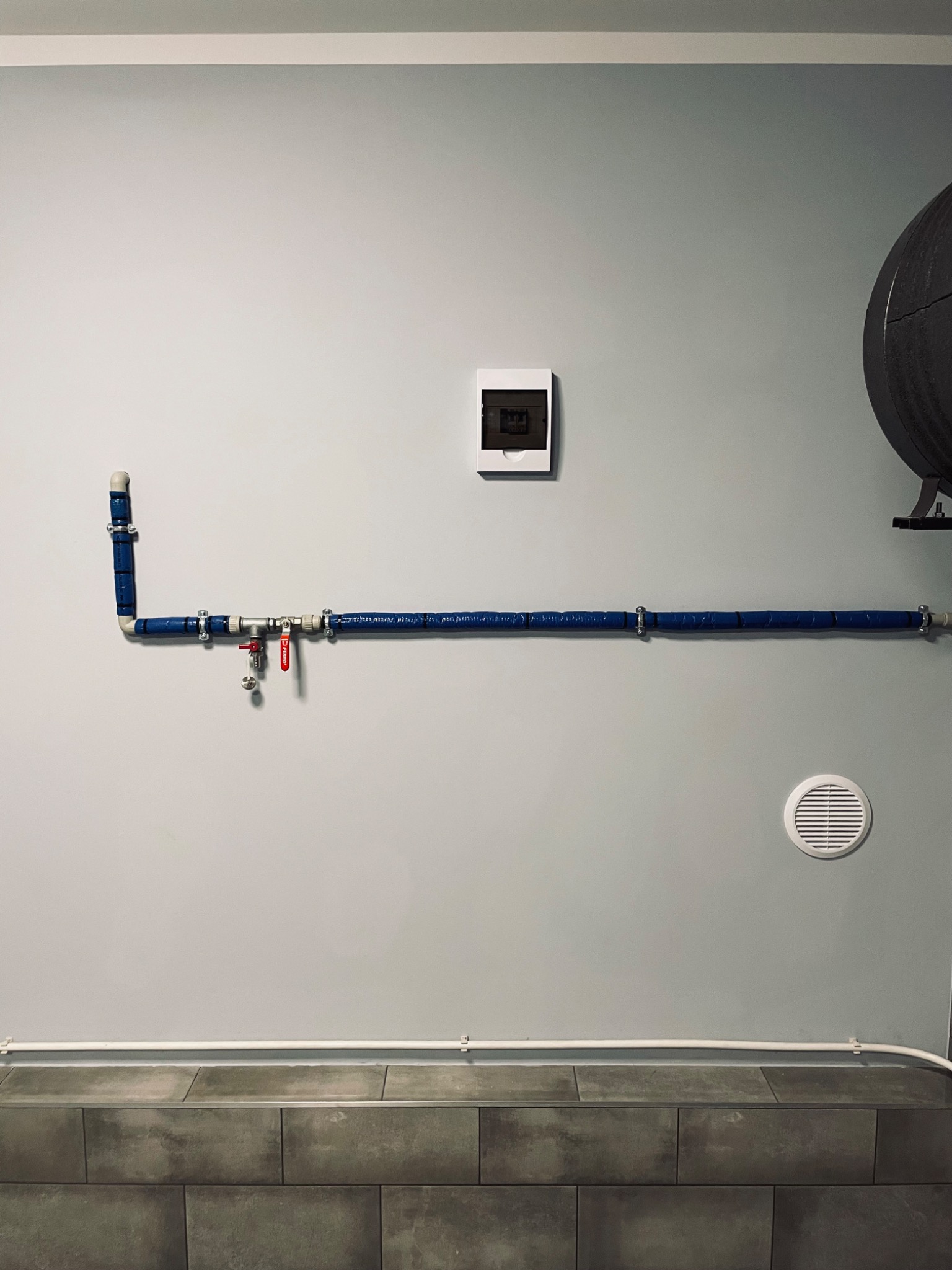 Niebieska, zaizolowana rura instalacji hydraulicznej z zaworem Ferro zamontowana na szarej ścianie nad ciemnymi płytkami, widoczny fragment zbiornika ciśnieniowego i kratka wentylacyjna.