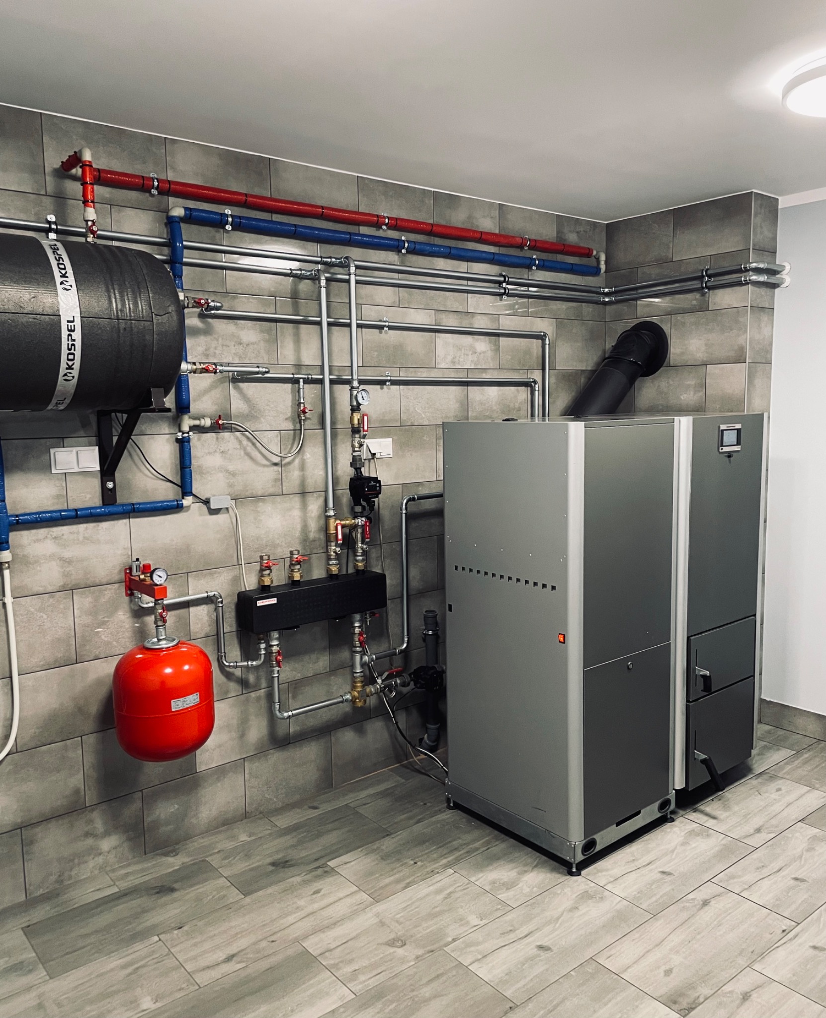 Zainstalowany piec grzewczy z zbiornikiem Kospel, orurowaniem w kolorach niebieskim i czerwonym oraz szarą płytką imitującą kamień na ścianach i podłodze.