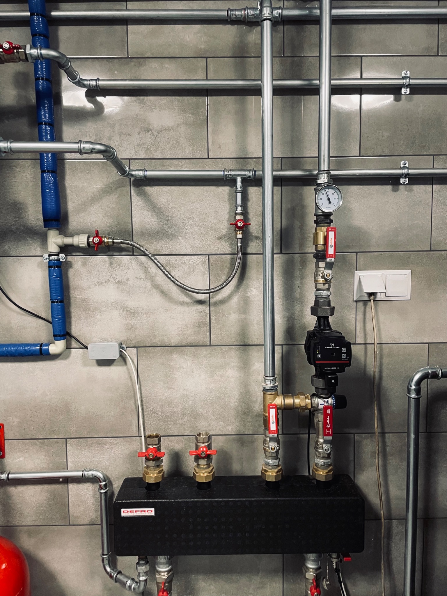 Rozbudowana instalacja grzewcza z licznymi rurami, zaworami, manometrem i pompą obiegową na tle ściany wyłożonej szarymi płytkami, widoczny czarny rozdzielacz DEFRO.