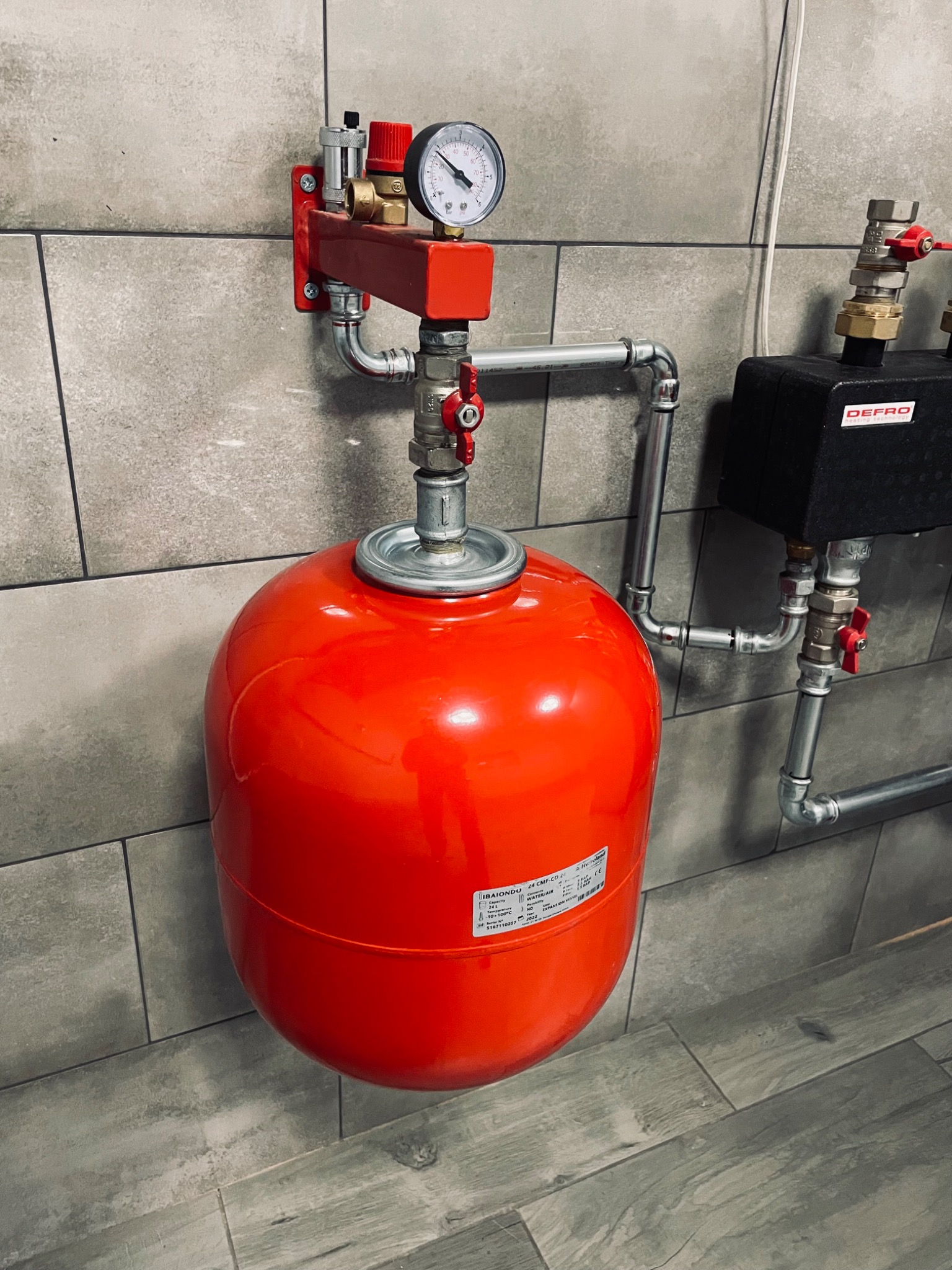 Czerwony zbiornik wyrównawczy ciśnienia z manometrem i zaworami, zamontowany na ścianie wyłożonej szarymi płytkami, z widocznymi rurami instalacji.