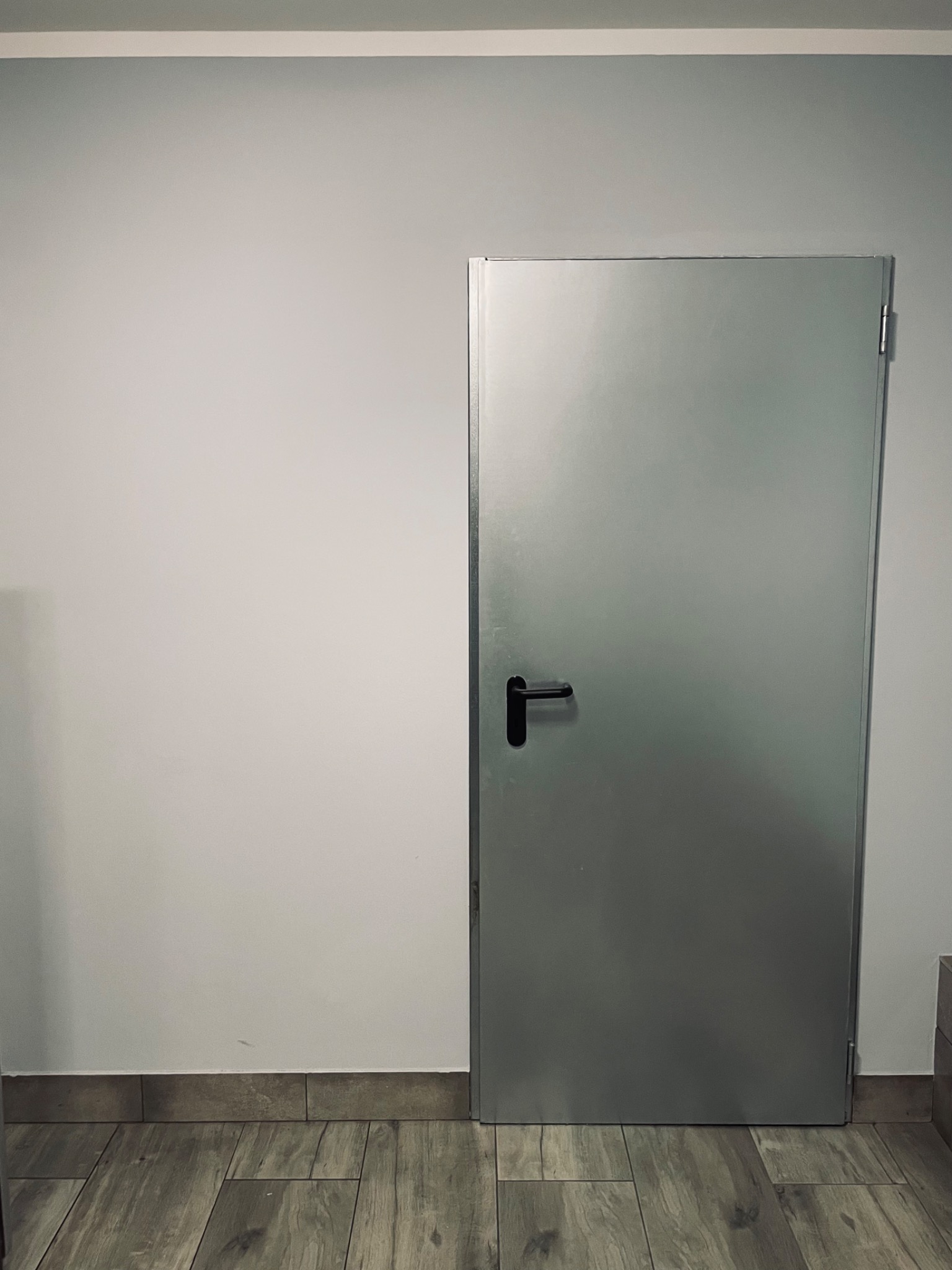 Zamknięte, metalowe drzwi z czarną klamką, w szarym pomieszczeniu z panelami podłogowymi imitującymi drewno.