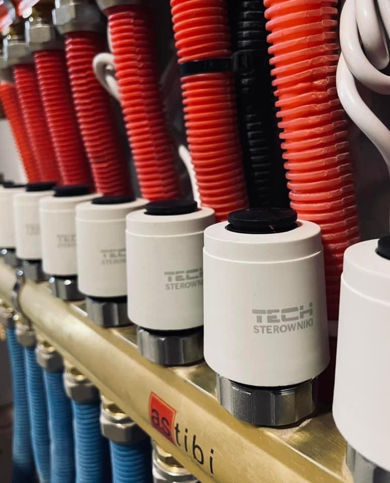 Rozdzielacz ogrzewania podłogowego z termostatami TECH, widoczne czerwone i niebieskie rury z tworzywa sztucznego.