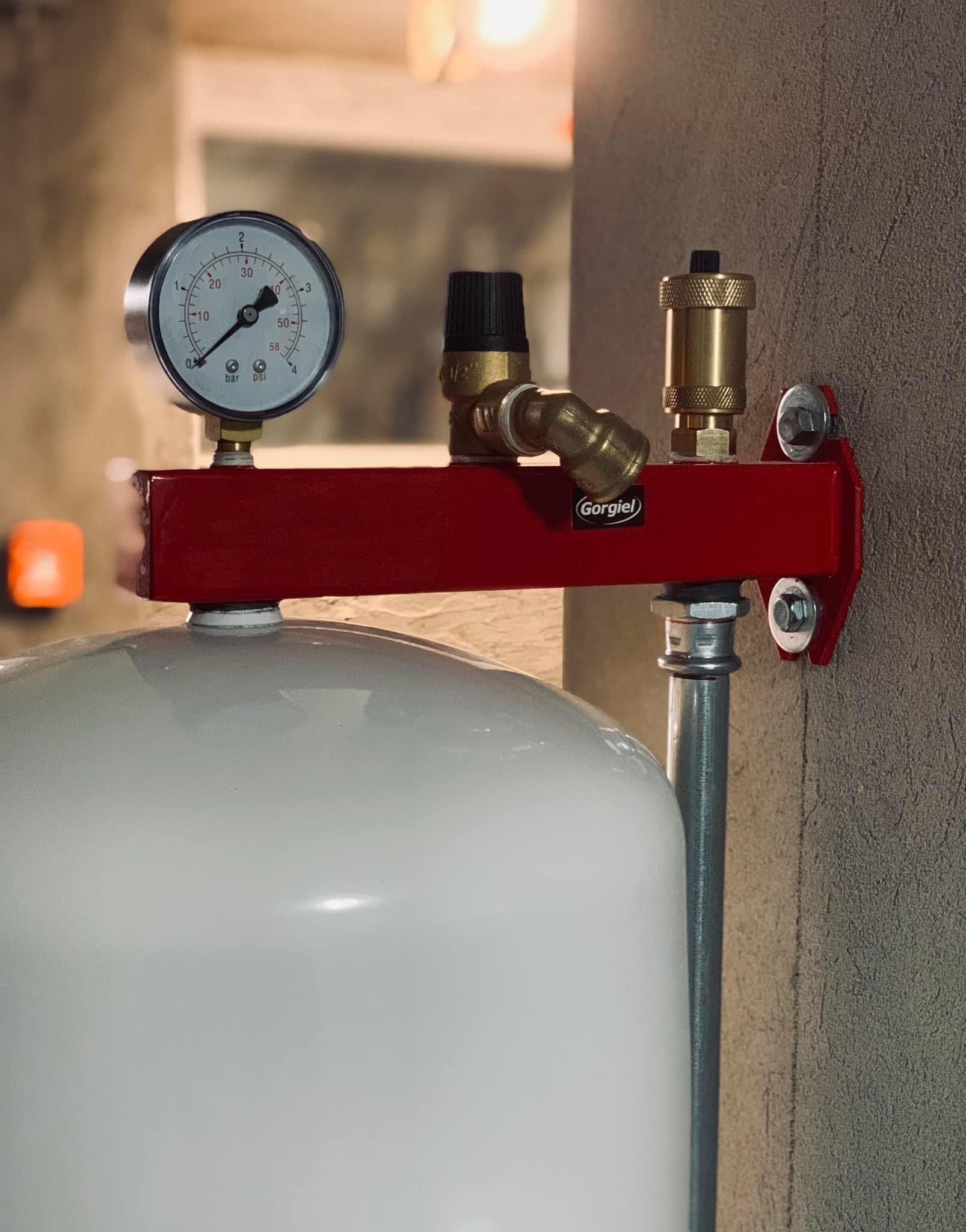 Biały zbiornik ciśnieniowy z czerwonym osprzętem, manometrem i zaworami, marki Gorgiel, zamontowany na szarej ścianie.