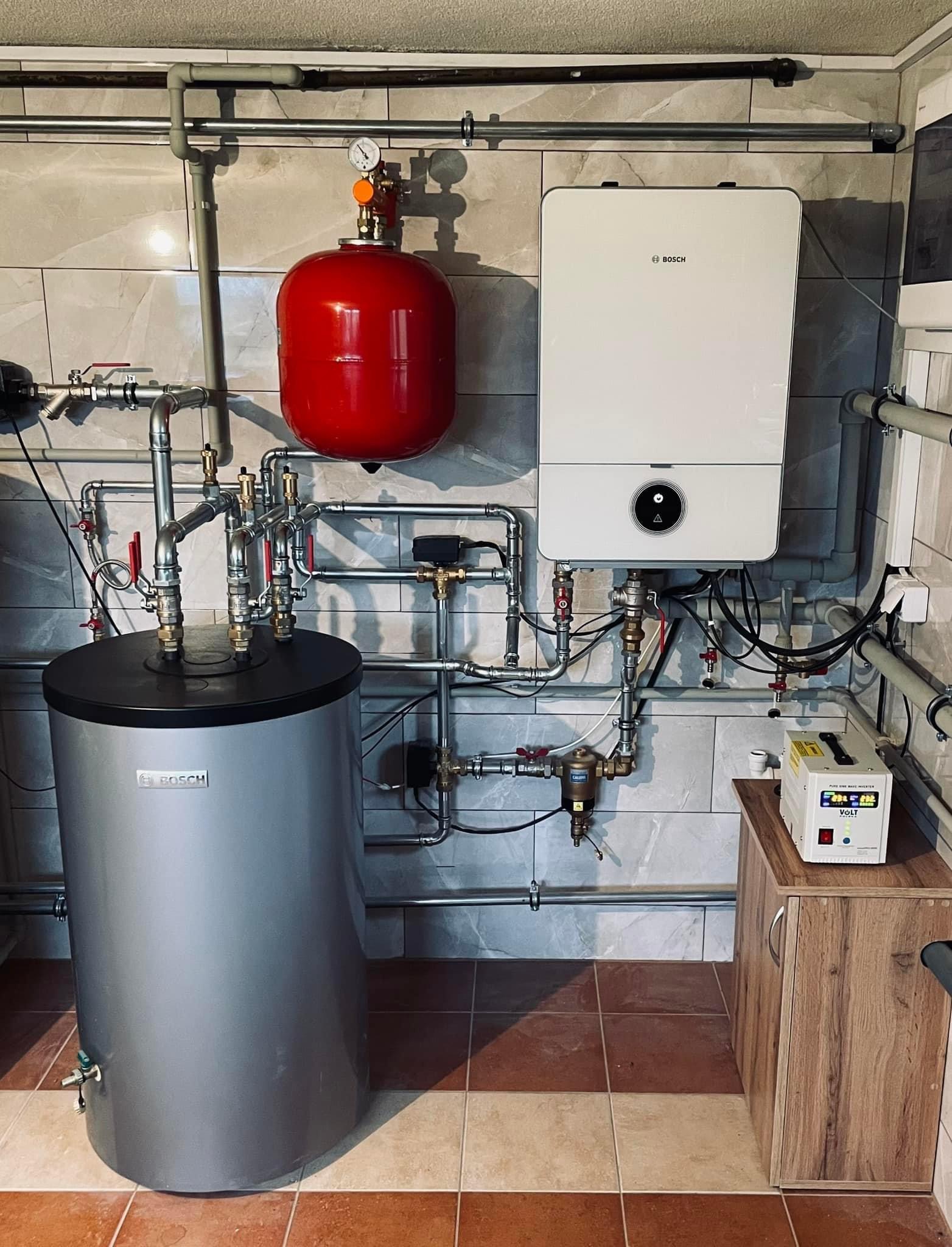 Kompletna instalacja kotła gazowego Bosch z zasobnikiem ciepłej wody, czerwonym naczyniem przeponowym i stabilizatorem napięcia Volt, zamontowana na tle płytek ceramicznych w odcieniach szarości...