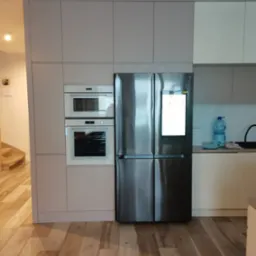 Jasnoszara zabudowa kuchenna z piekarnikiem, mikrofalówką i lodówką z ekranem dotykowym, na drewnianej podłodze.