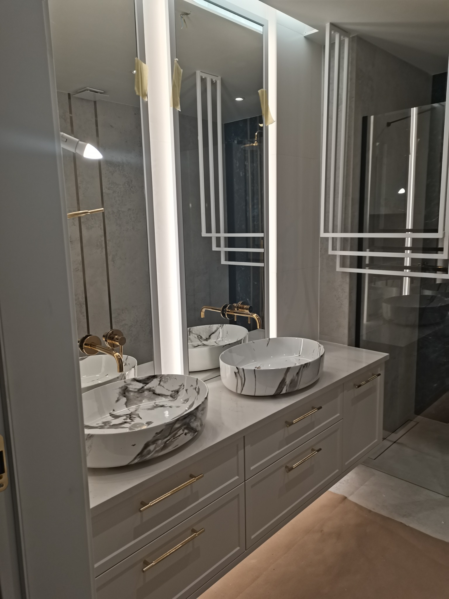 Nowoczesna łazienka z dwoma umywalkami w marmurkowy wzór, złotą armaturą i podświetlanymi lustrami, szafka z szufladami w kolorze szarym.