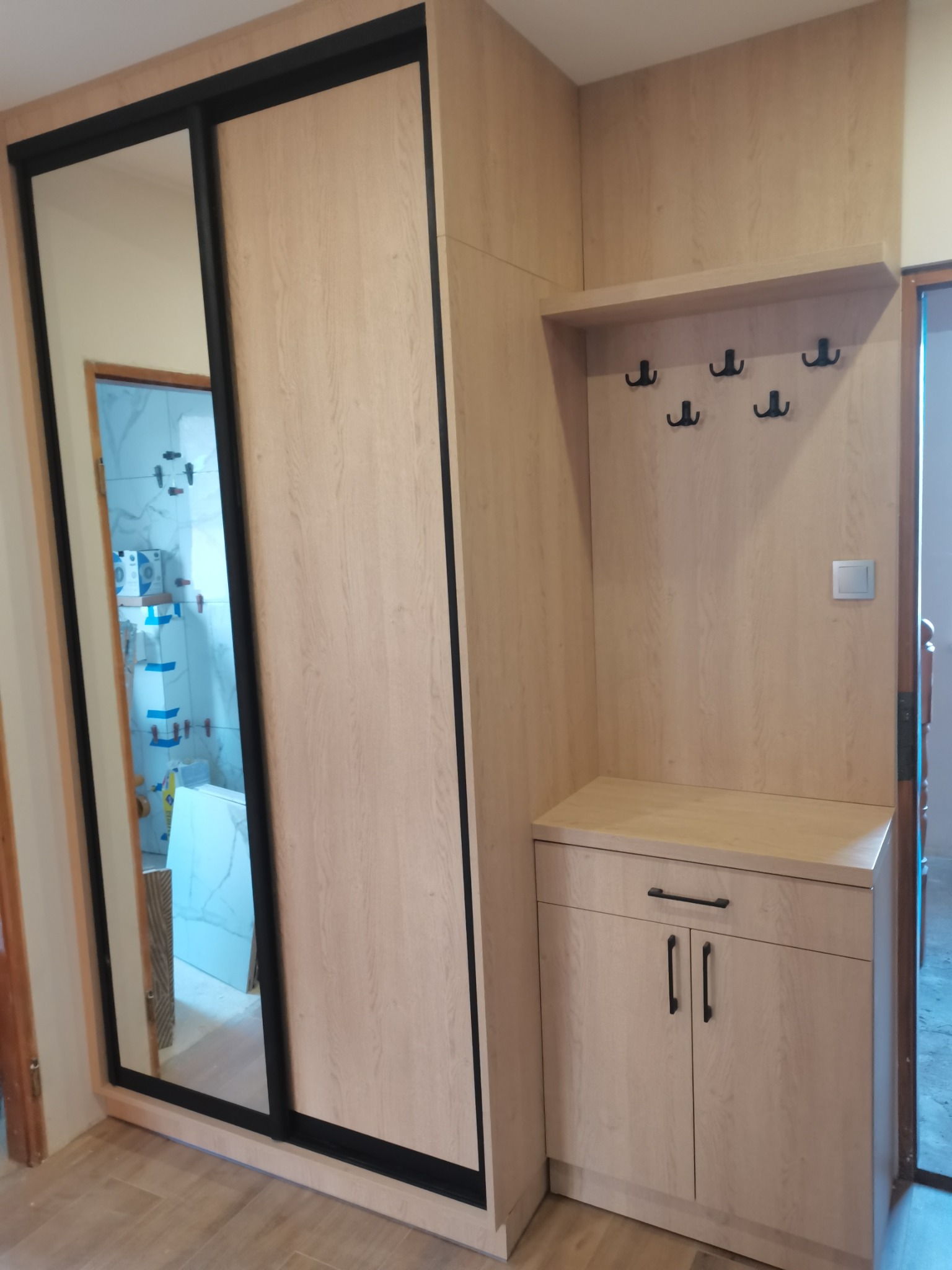 Jasna szafa przesuwna z czarnymi obramowaniami drzwi i lustrem, obok zabudowa z wieszakami, szafką i półką, całość w odcieniach jasnego drewna.