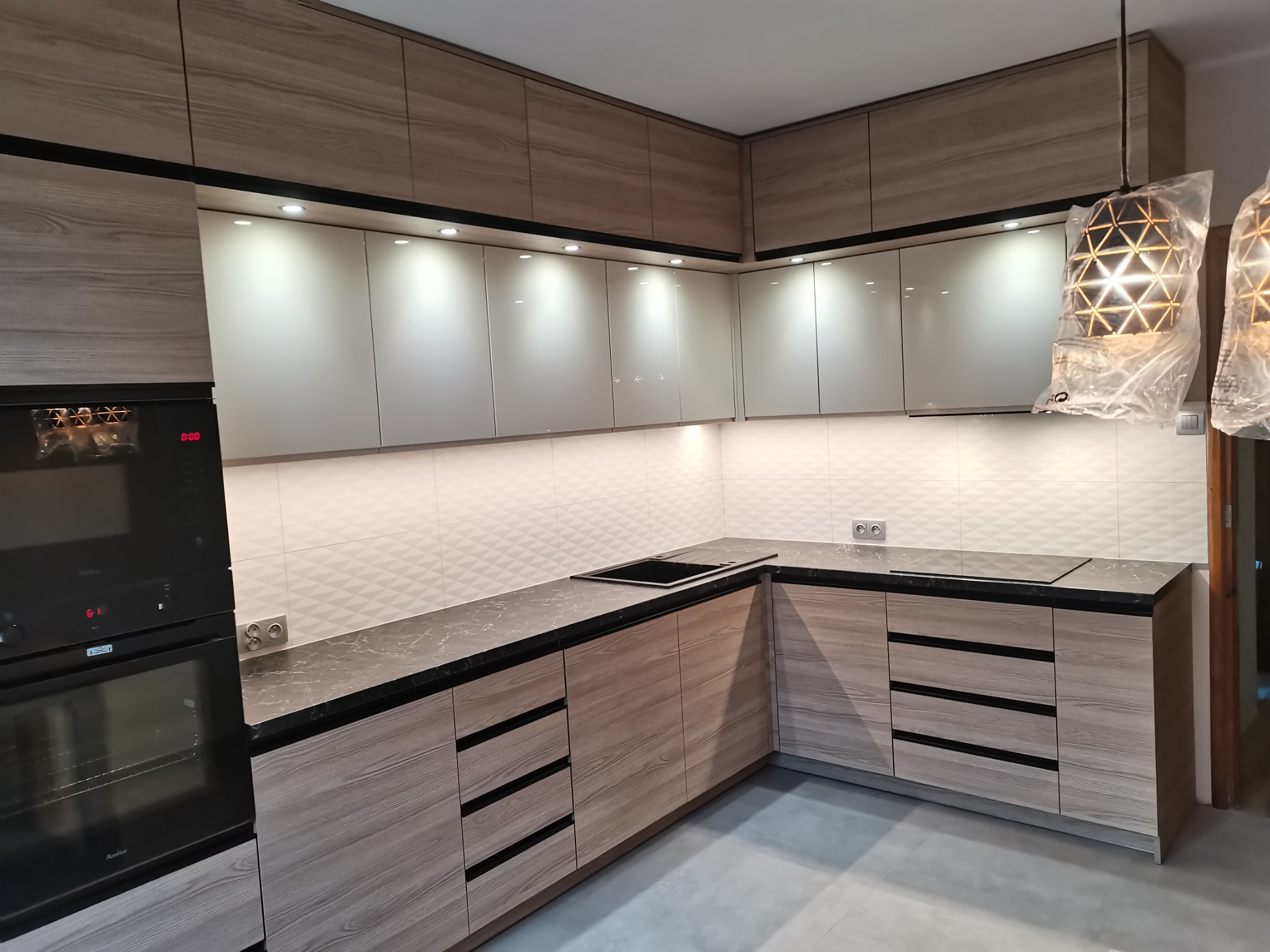 Nowoczesna kuchnia z drewnopodobnymi frontami szafek, szarym blatem i białymi kafelkami na ścianie, oświetlona punktowymi lampami podszafkowymi i wiszącymi lampami w folii ochronnej.