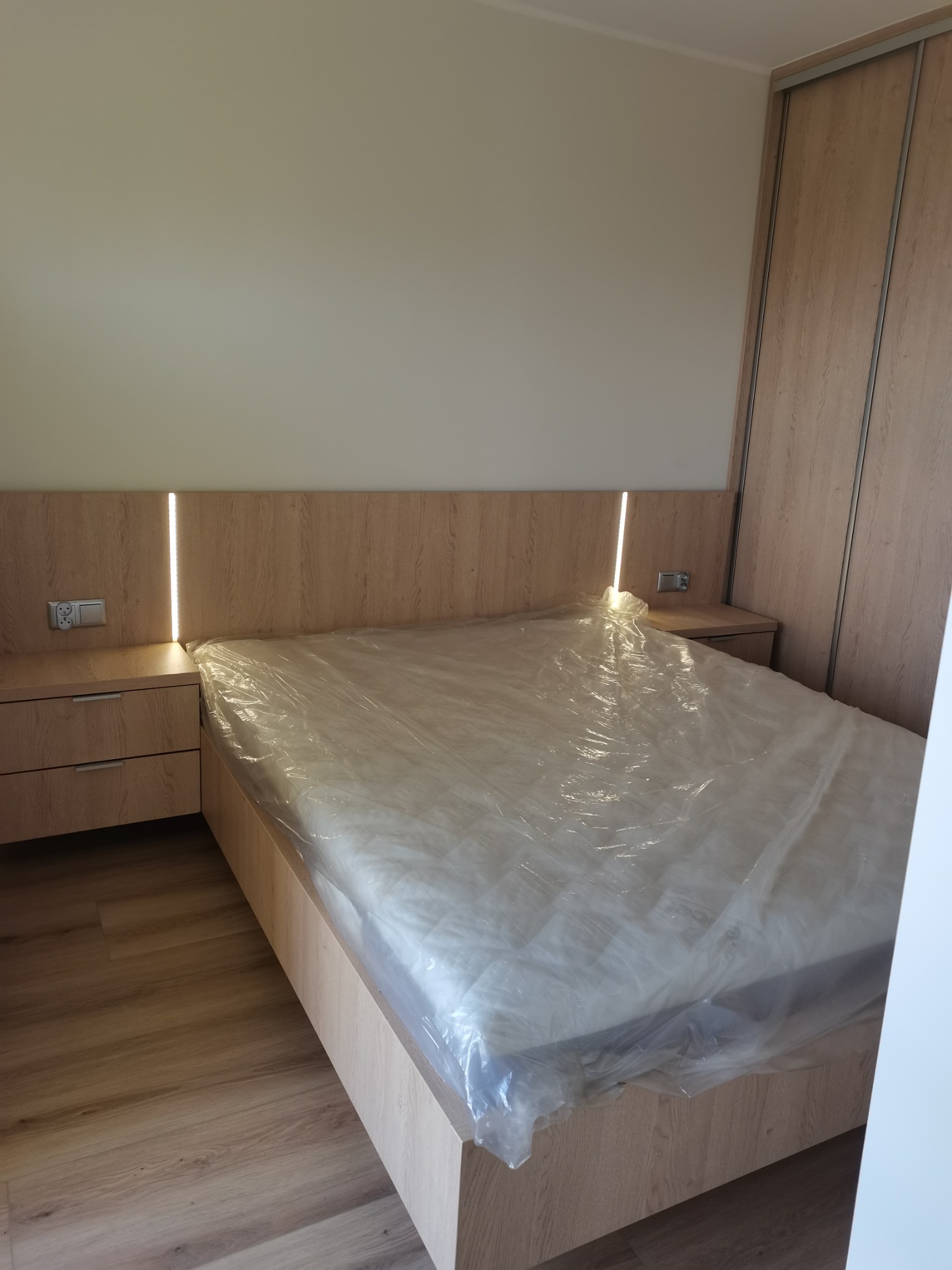 Drewniane łóżko z podświetlanym zagłówkiem i szafkami nocnymi, materac zabezpieczony folią, obok szafa przesuwna z drewnianymi frontami.