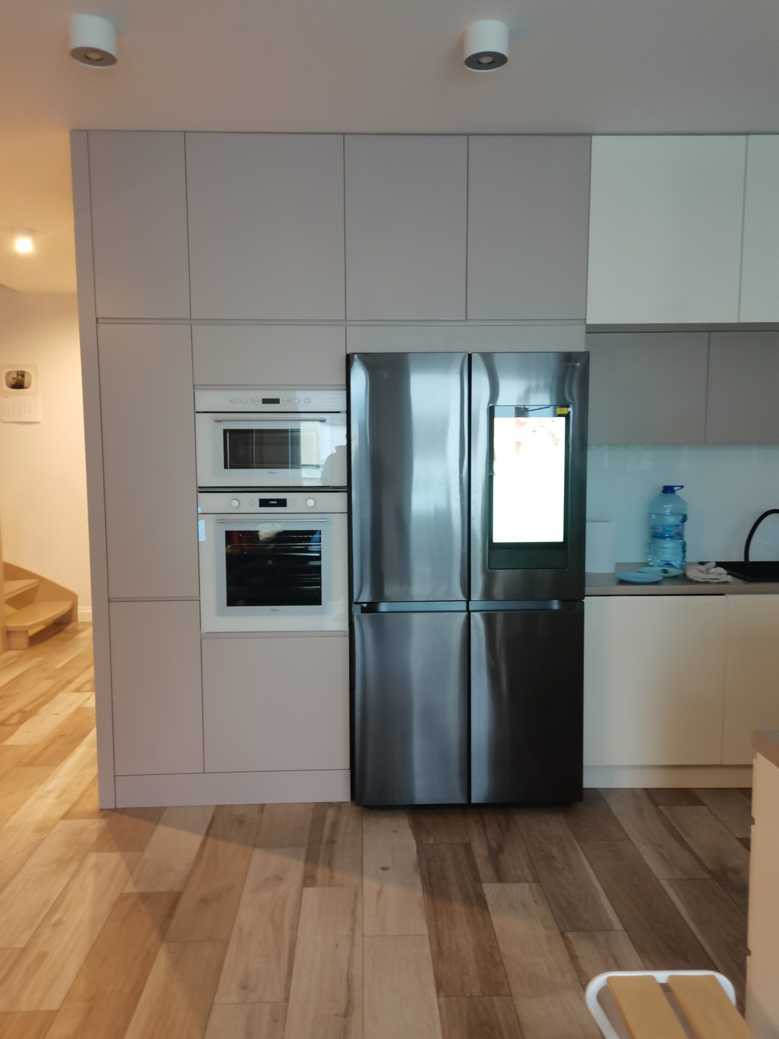 Jasnoszara zabudowa kuchenna z piekarnikiem, mikrofalówką i lodówką z ekranem dotykowym, na drewnianej podłodze.