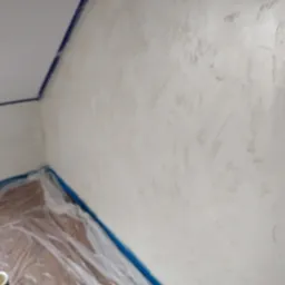 Ściana po nałożeniu dekoracyjnej, jasnej struktury imitującej kamień, zabezpieczona folią podłoga, widoczne niebieskie taśmy malarskie.