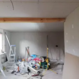 Wnętrze w trakcie remontu: ściany obłożone płytami gipsowo-kartonowymi, widoczna drewniana belka stropowa, narzędzia i materiały budowlane w nieładzie.