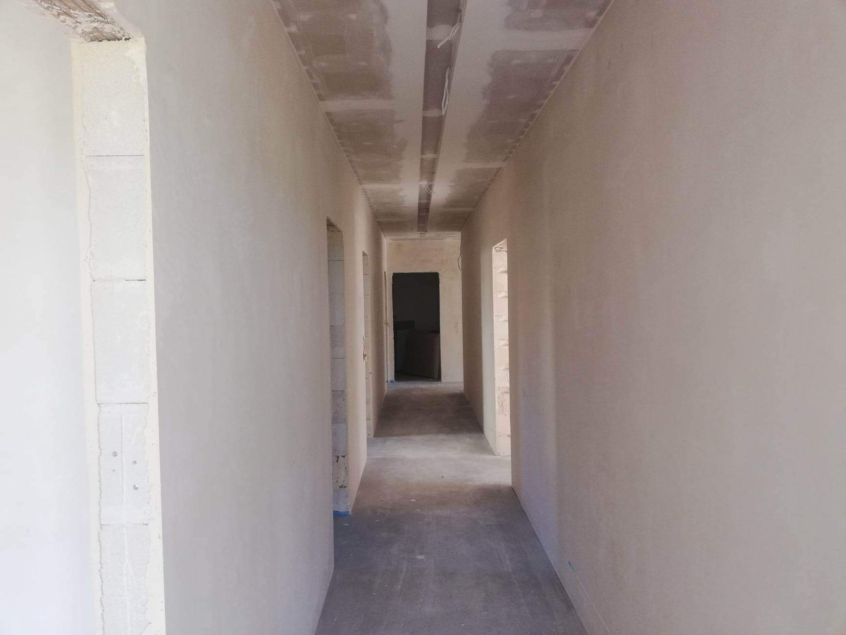 Korytarz w trakcie prac wykończeniowych; ściany pokryte gładzią, sufit z zamontowaną płytą kartonowo-gipsową, widoczne surowe otwory drzwiowe.