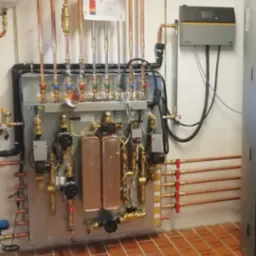 Rozbudowana instalacja centralnego ogrzewania z miedzianymi rurami, zaworami i sterownikiem elektronicznym zamontowana na ścianie z terakotową podłogą.