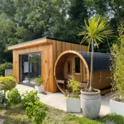 Nowoczesny drewniany domek z bali z płaskim dachem i przeszklonymi drzwiami przesuwnymi, obok beczka sauny z czarnym dachem, otoczone zielenią i roślinami w donicach na jasnym brukowanym tarasie.