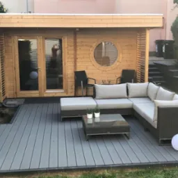 Nowoczesna drewniana konstrukcja z okrągłym oknem, wyposażona w meble ogrodowe na szarym tarasie kompozytowym, oświetlona dekoracyjnymi kulami.