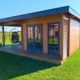 Nowoczesna, drewniana konstrukcja z płaskim dachem i niebieskimi drzwiami, otoczona zielonym trawnikiem, z wiszącym fotelem na tarasie.