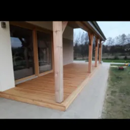 Zadaszony taras z jasnego drewna przylegający do domu z dużym oknem tarasowym, widoczne drewniane słupy nośne i deski podłogowe.