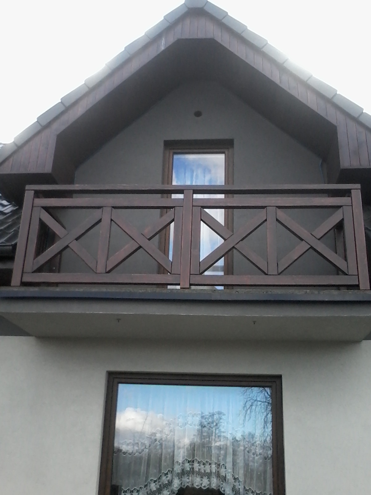 Ciemnobrązowa, drewniana balustrada balkonowa z krzyżowym wzorem, widoczna na tle okna poddasza w domu w Połomi. W oknach odbija się błękitne niebo.