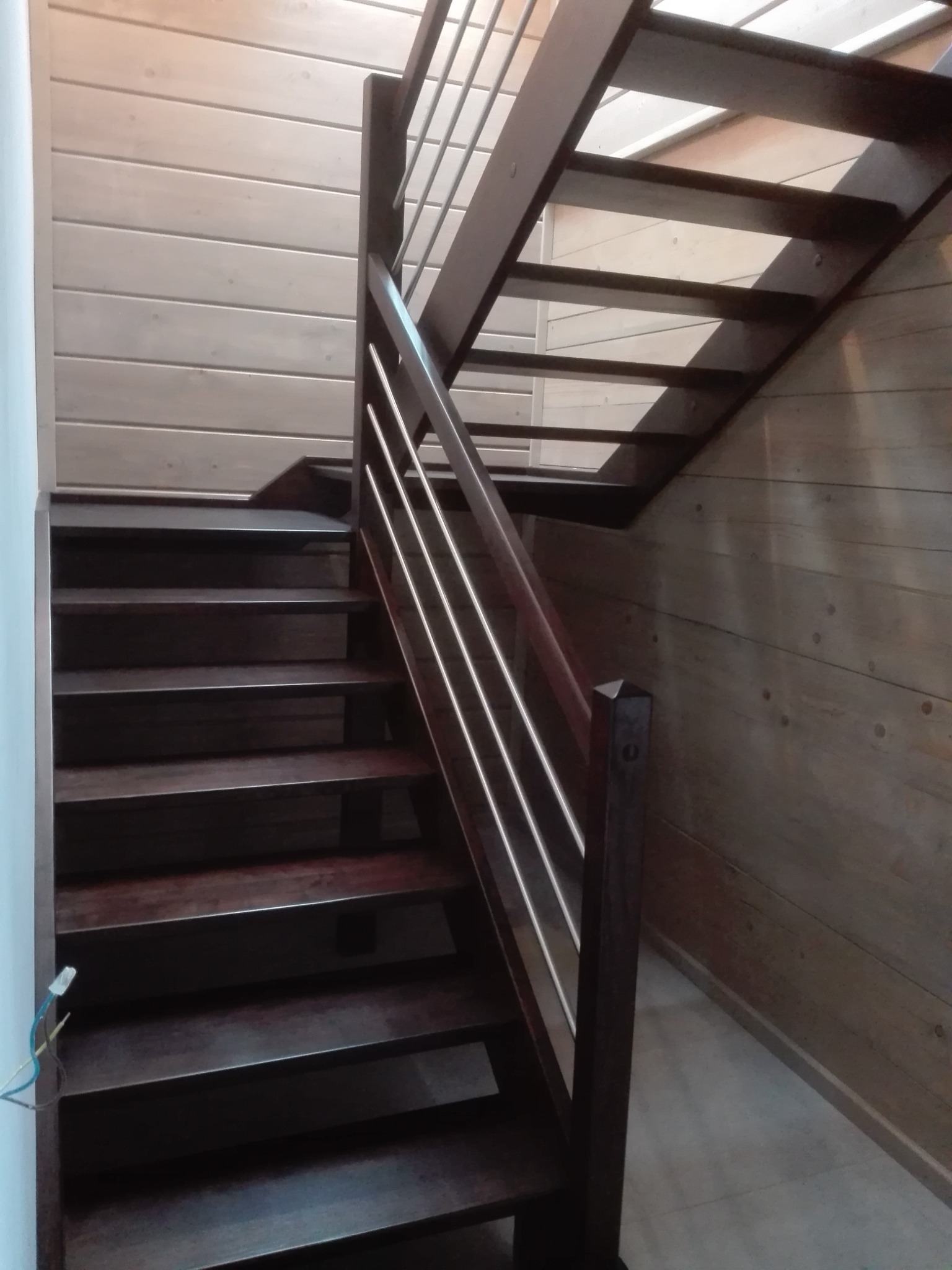 Ciemne, drewniane stopnie schodów z metalową balustradą w jasnym wnętrzu wykończonym boazerią.