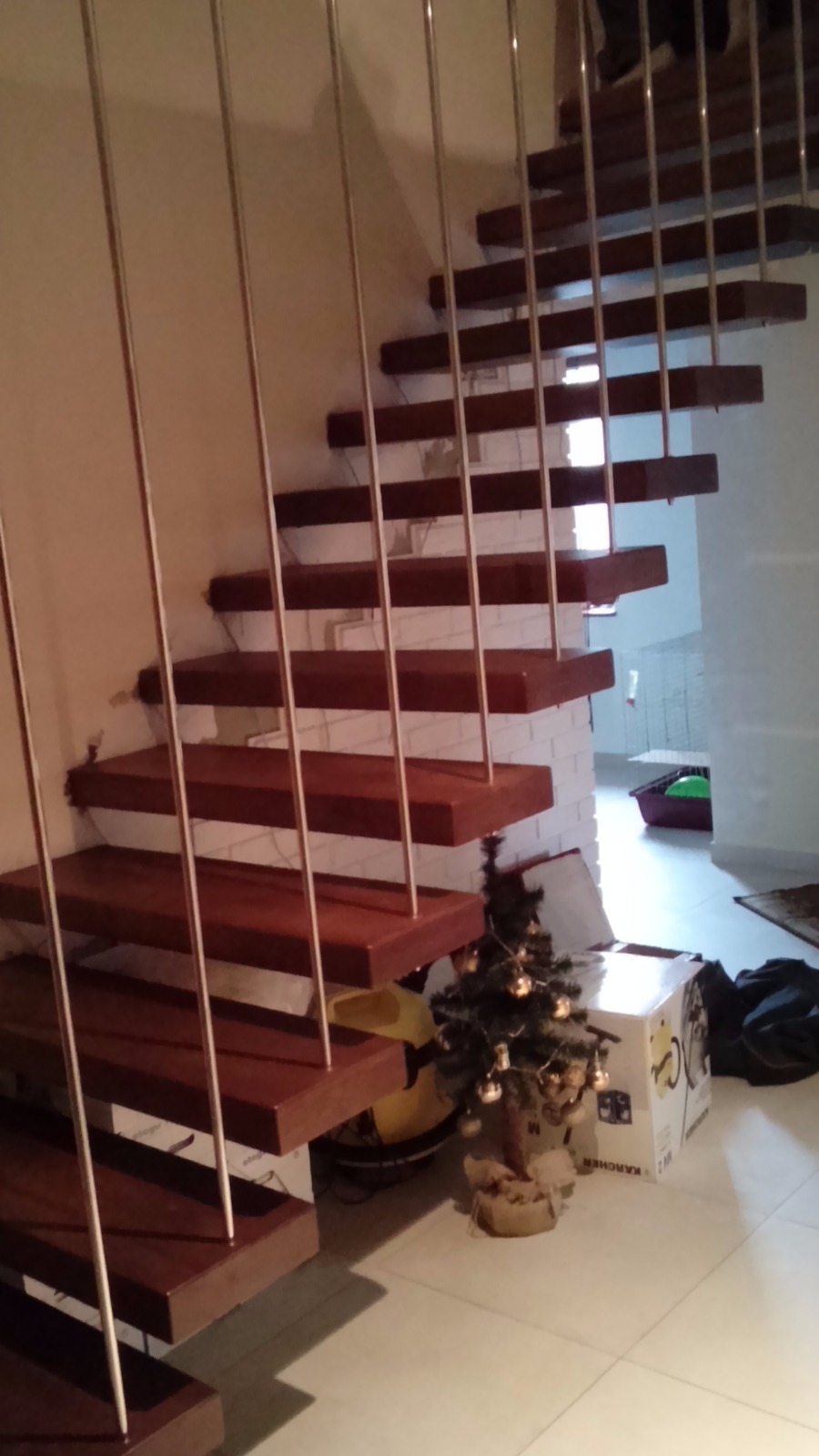 Nowoczesne schody z drewnianymi stopniami i metalowymi barierkami, widziane z dołu, z małą choinką i kartonem Karcher u podstawy.