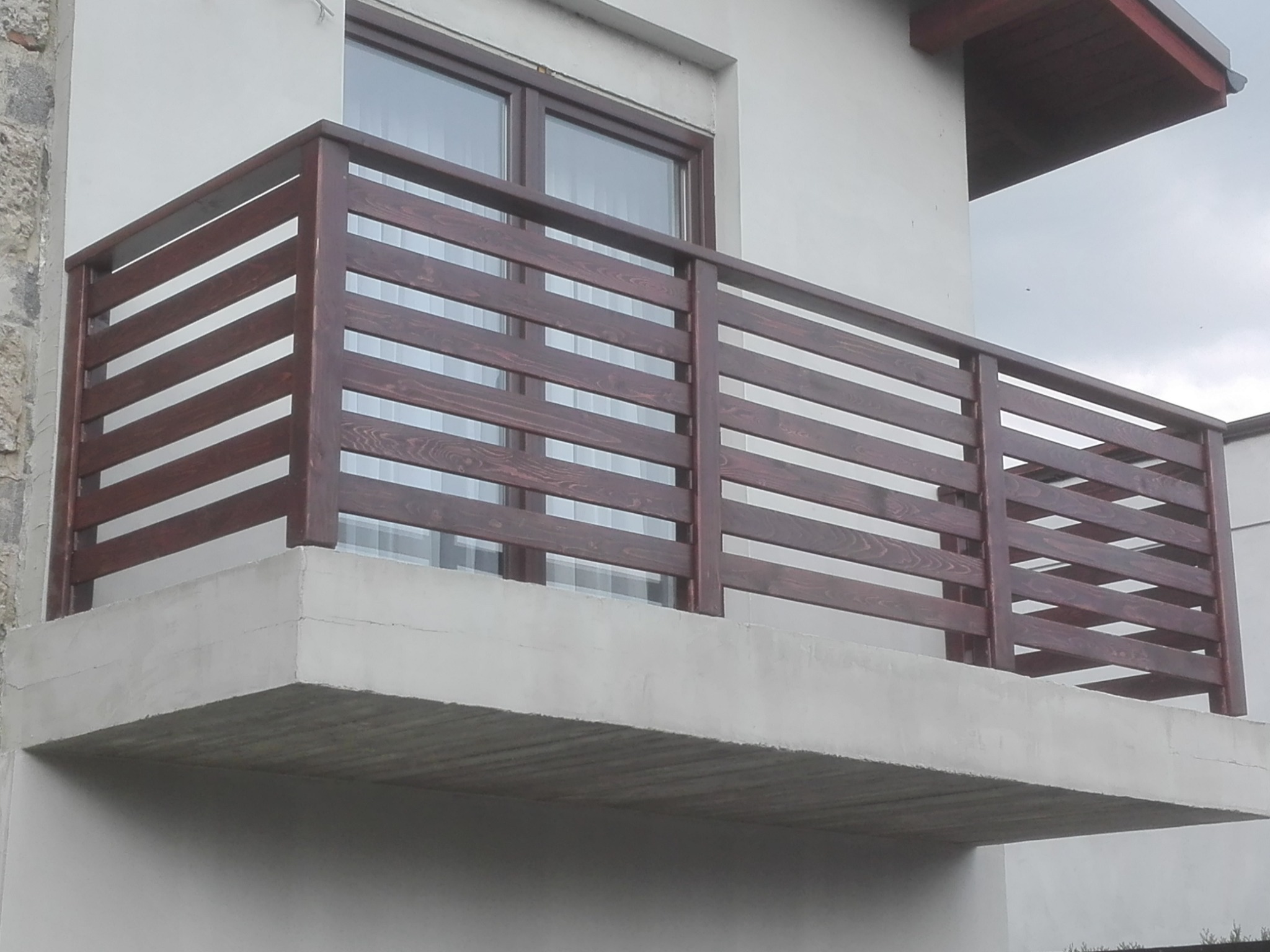 Drewniana balustrada balkonowa z poziomych desek o ciemnobrązowym odcieniu, zamontowana na betonowej płycie balkonu, widok na okno i część dachu budynku.