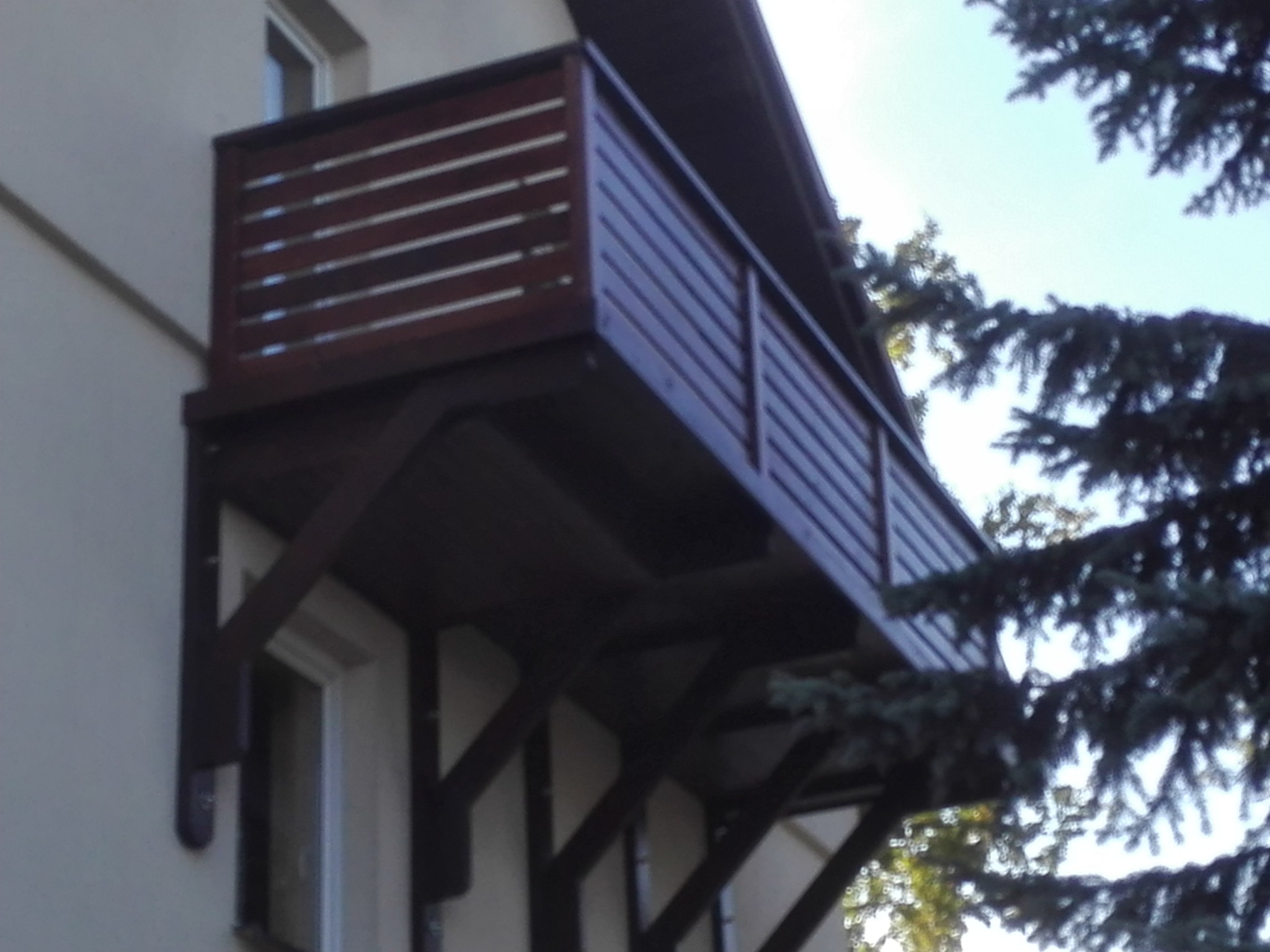 Drewniany balkon z poziomymi deskami balustrady, zamontowany na wspornikach, widoczny z dołu na tle jasnej ściany budynku i iglastego drzewa.