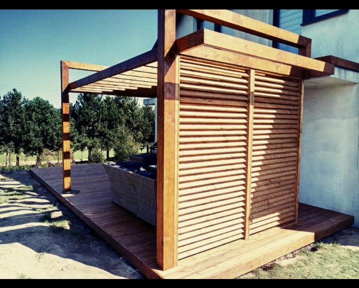 Drewniana konstrukcja z żaluzjami i zadaszeniem na tarasie, z widoczną sofą i zielenią w tle, oświetlona popołudniowym słońcem.