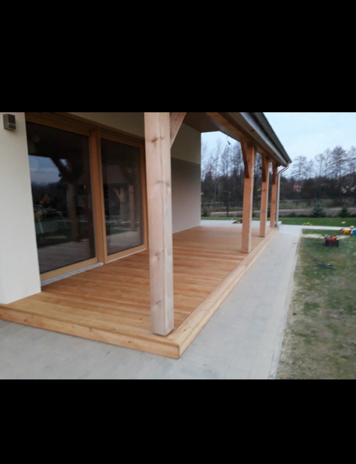 Zadaszony taras z jasnego drewna przylegający do domu z dużym oknem tarasowym, widoczne drewniane słupy nośne i deski podłogowe.