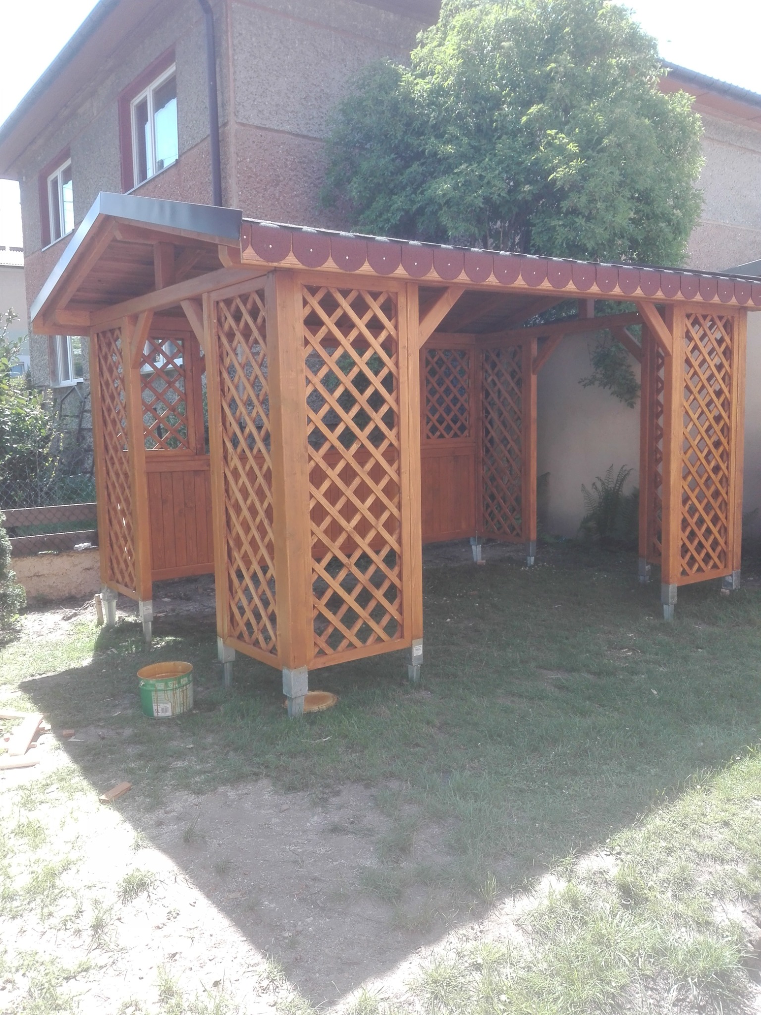 Nowo wybudowana, drewniana altana z kratownicowymi ścianami i czerwoną dachówką, ustawiona na trawniku przed domem, z widocznym wiaderkiem po farbie.